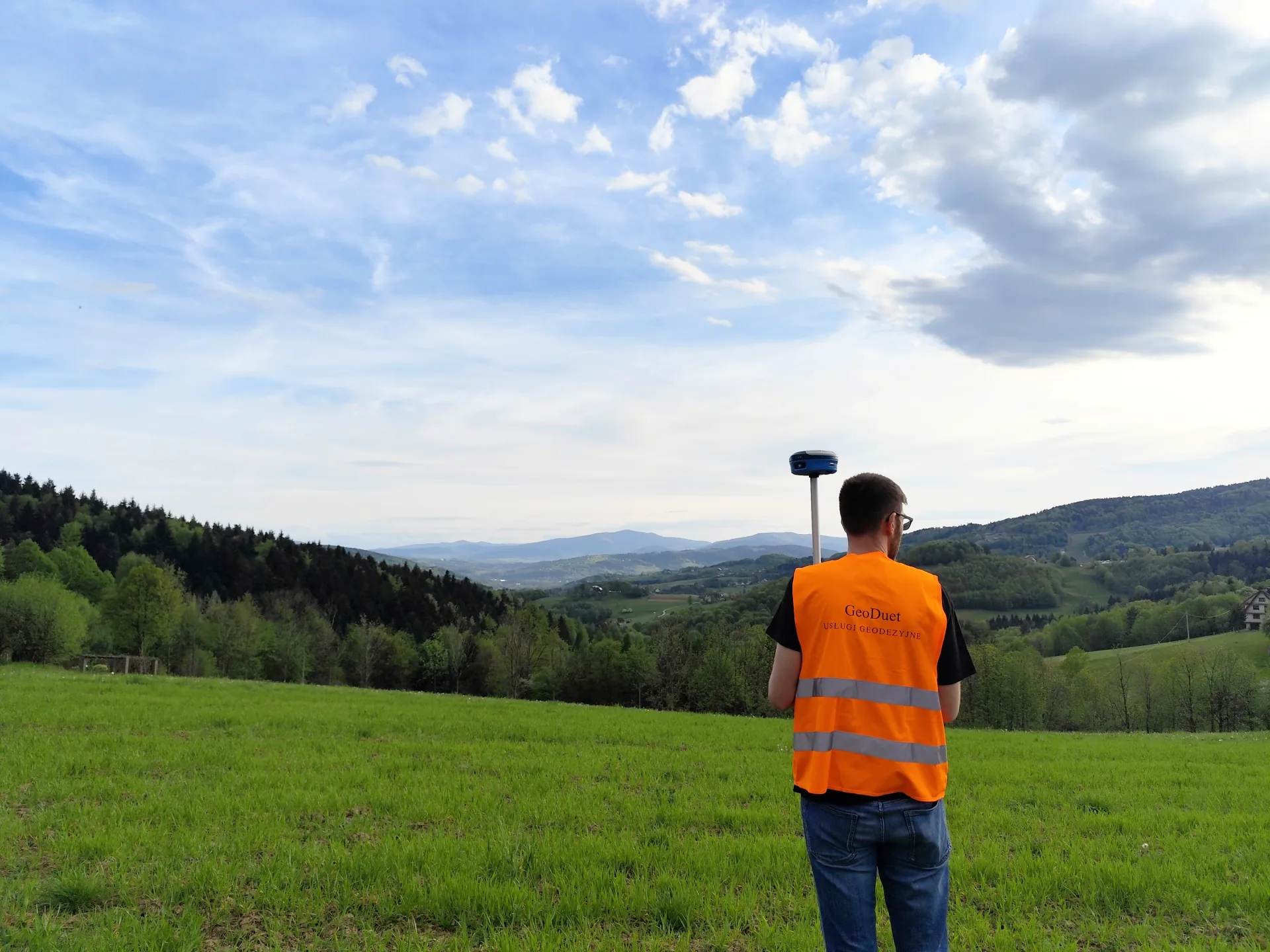 Geodeta podczas pomiaru odbiornikiem GNSS na tle zielonego, pagórkowatego terenu
